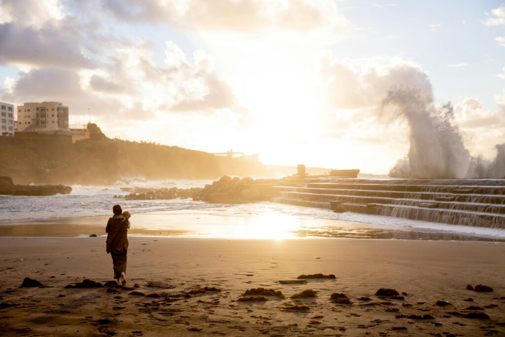 A parent carrying a child walks on the beach at sunrise, with dramatic waves crashing in the background.