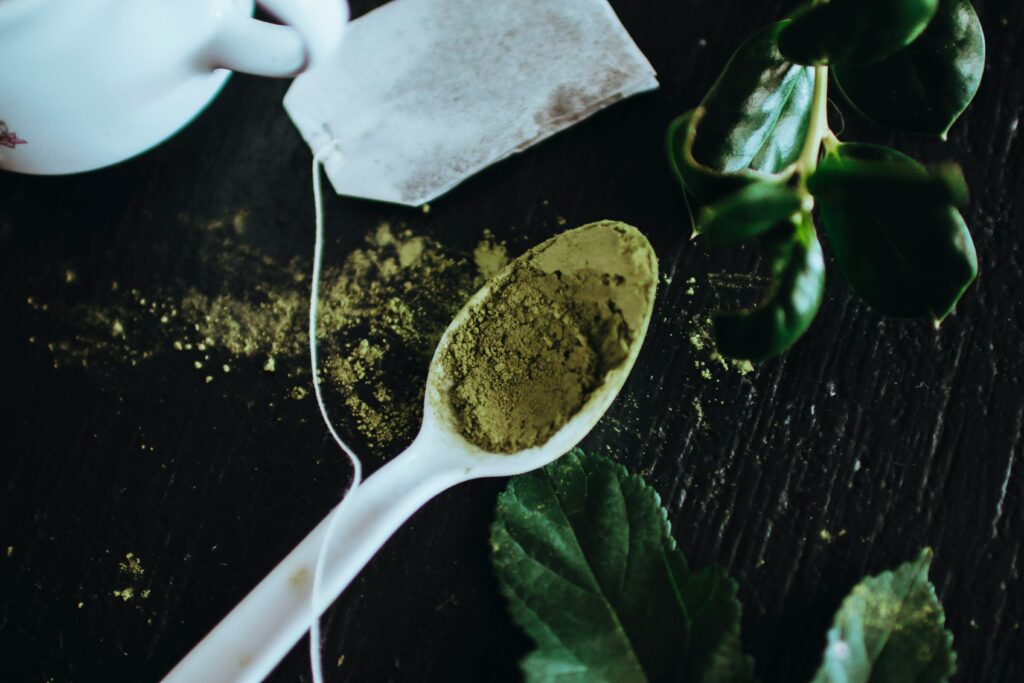 Close-up of matcha powder on a spoon with tea bag and green leaves on a dark table.