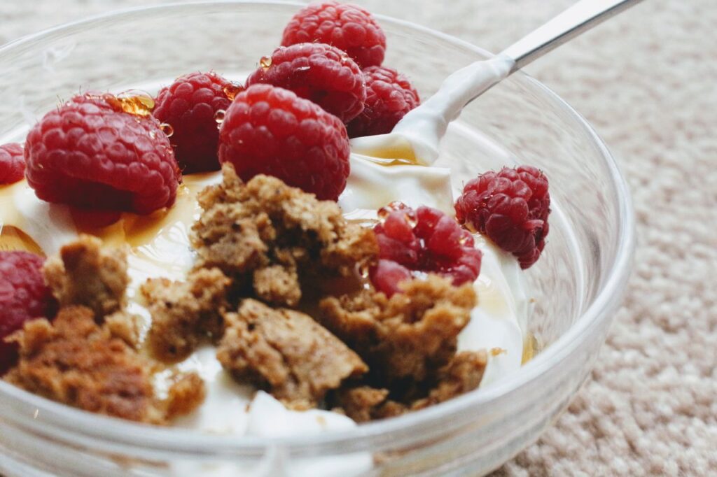 From above closeup of breakfast with tasty natural yogurt decorated with crumbled biscuit and whole ripe raspberries with caramel syrup in glass bowl with spoon on soft plaid in morning
